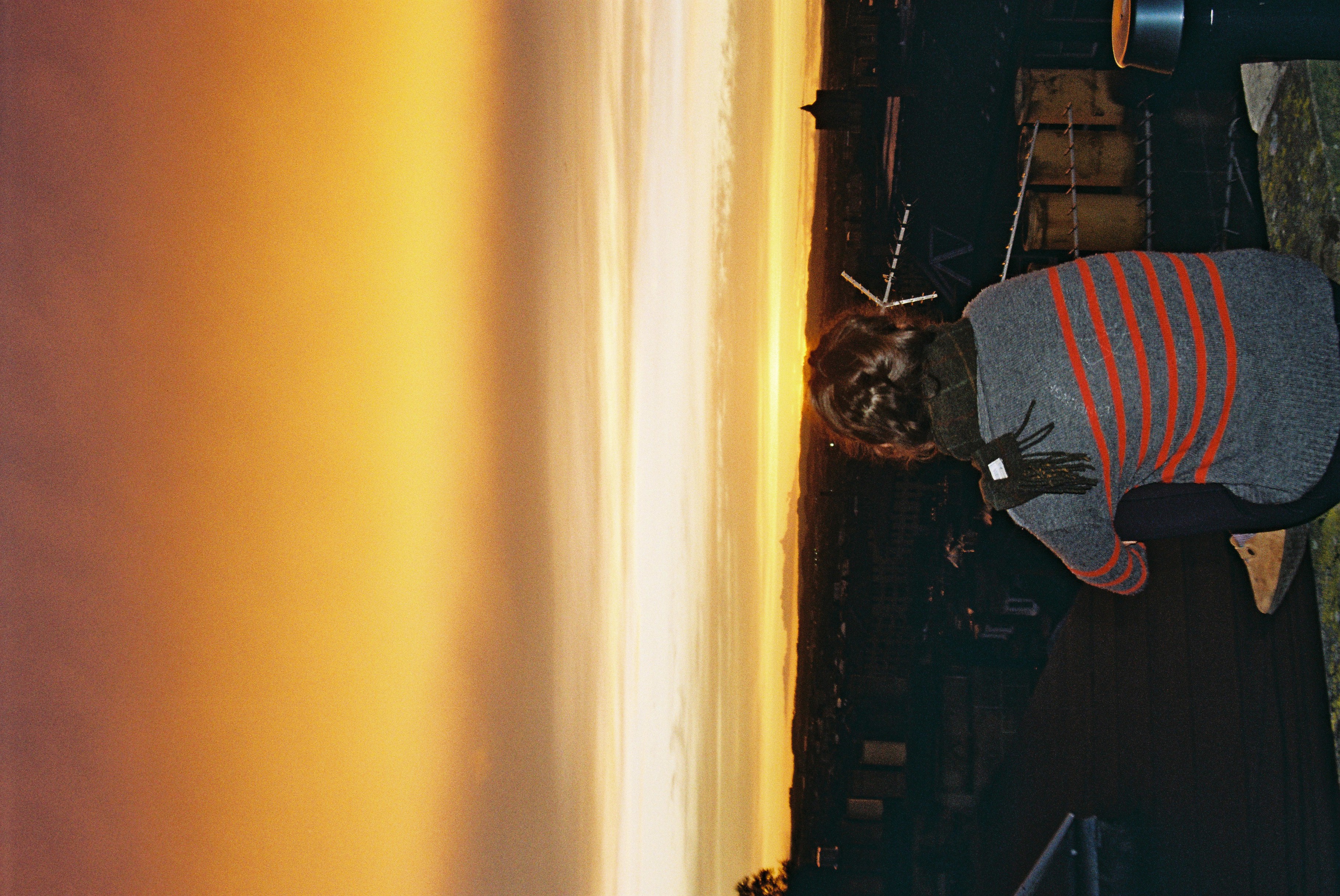 A girl sits on the roof with her back to the camera looking at the sunset
