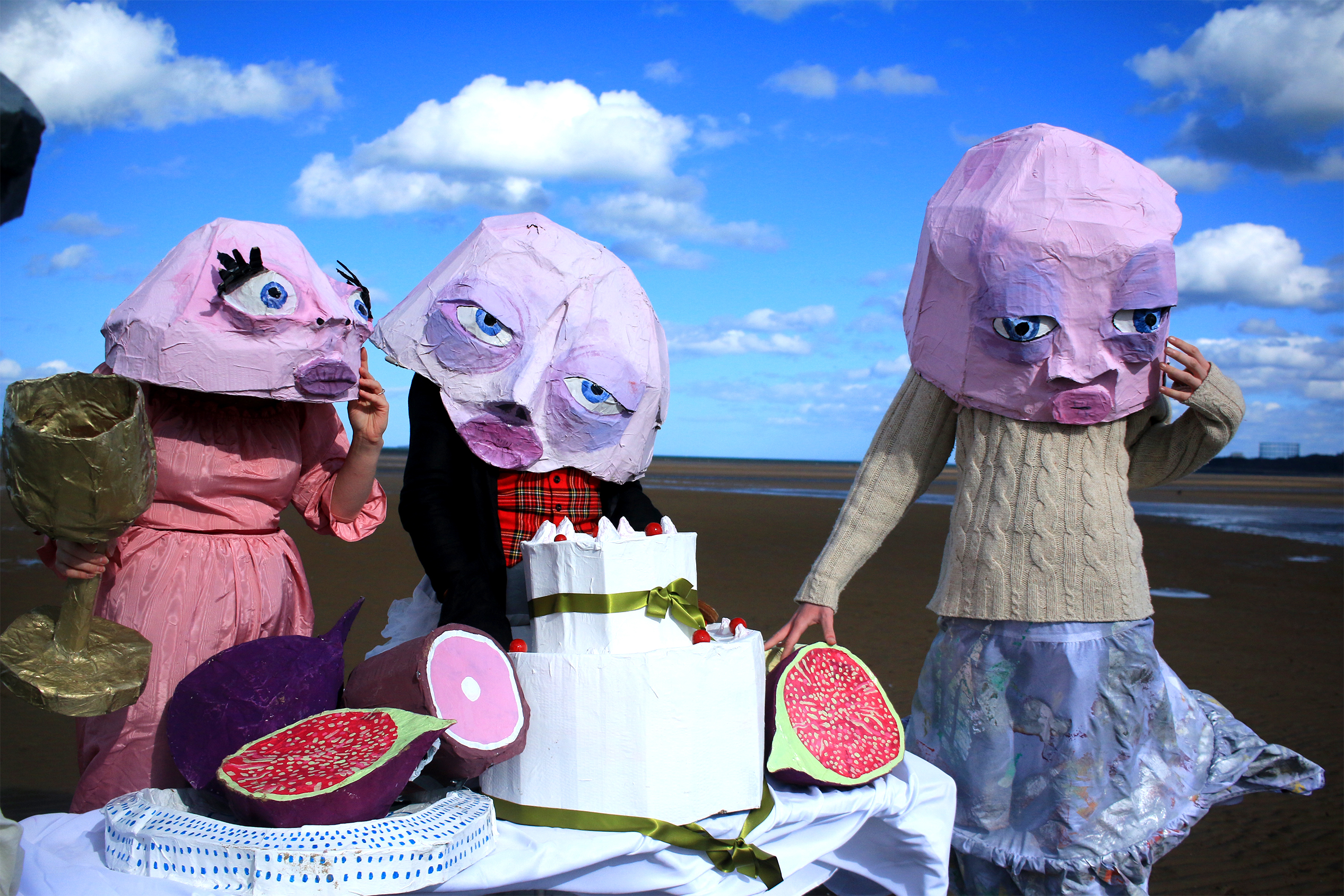 Figures stood around a table of cardboard food on the beach with pink papier-mâché masks on.