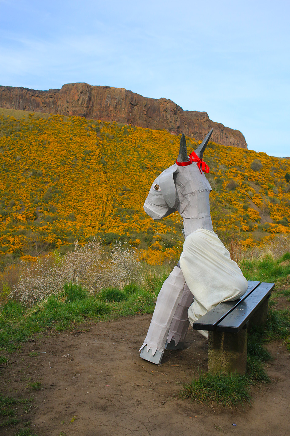 Figure in papier-mâché goat costume sat on a bench, looking at yellow flowers.