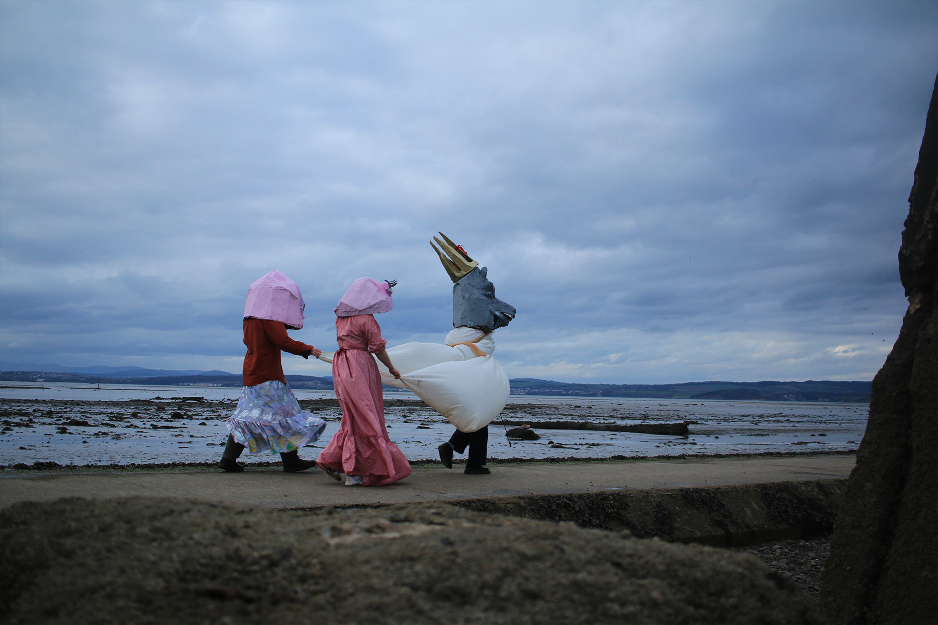 Two figures with papier-mâché masks holding the cloak of another figure wearing a papier-mâché goat head and crown as they walk across a concrete causeway.