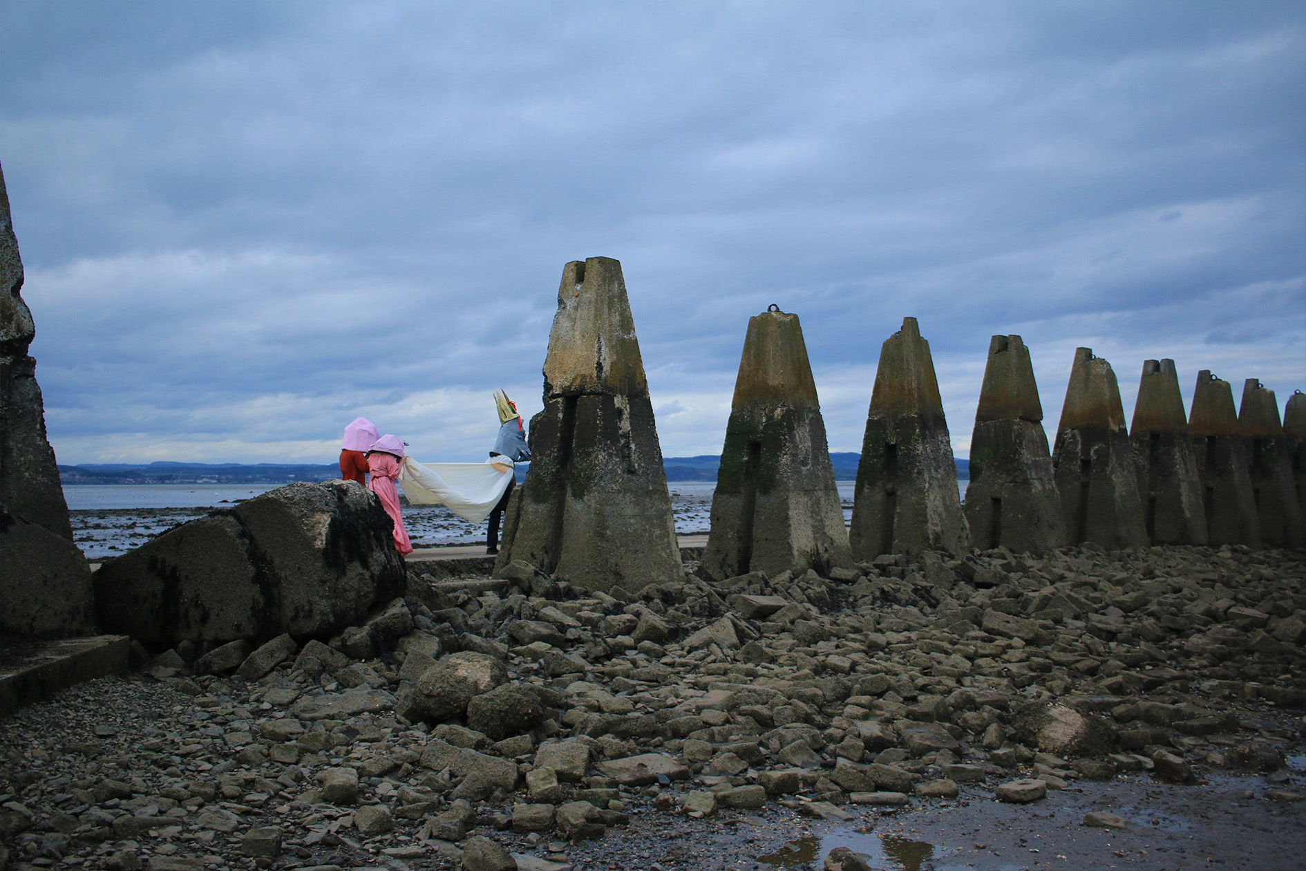 Two figures with papier-mâché masks holding the cloak of another figure wearing a papier-mâché goat head and crown as they walk across a concrete causeway.