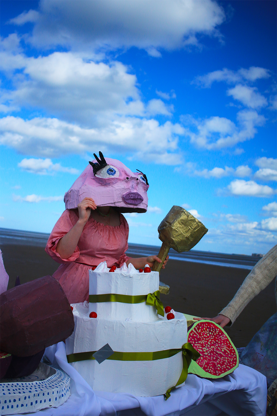 One figure in a pink papier-mâché mask stood next to a table of card-board food on the beach.