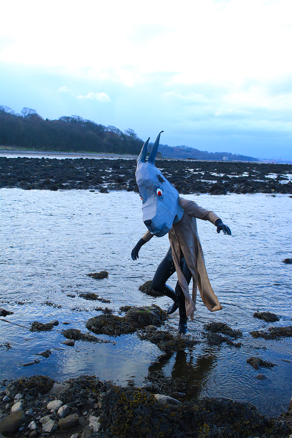 Figure with a papier-mâché goat mask walking out of the water at the beach.